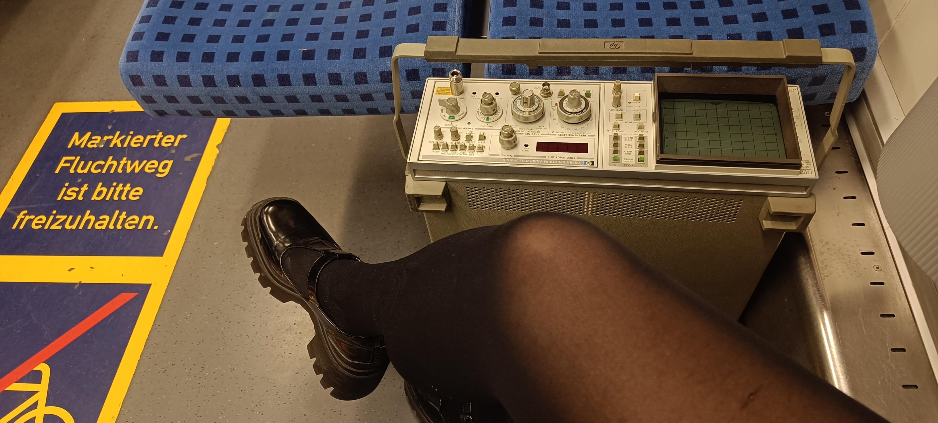 Ein großes eckiges Messgerät in einer S-Bahn auf dem Boden, daneben die überschlagenen Beine von Chrissy Ein großes eckiges Messgerät in einer S-Bahn auf dem Boden, daneben die überschlagenen Beine von Chrissy