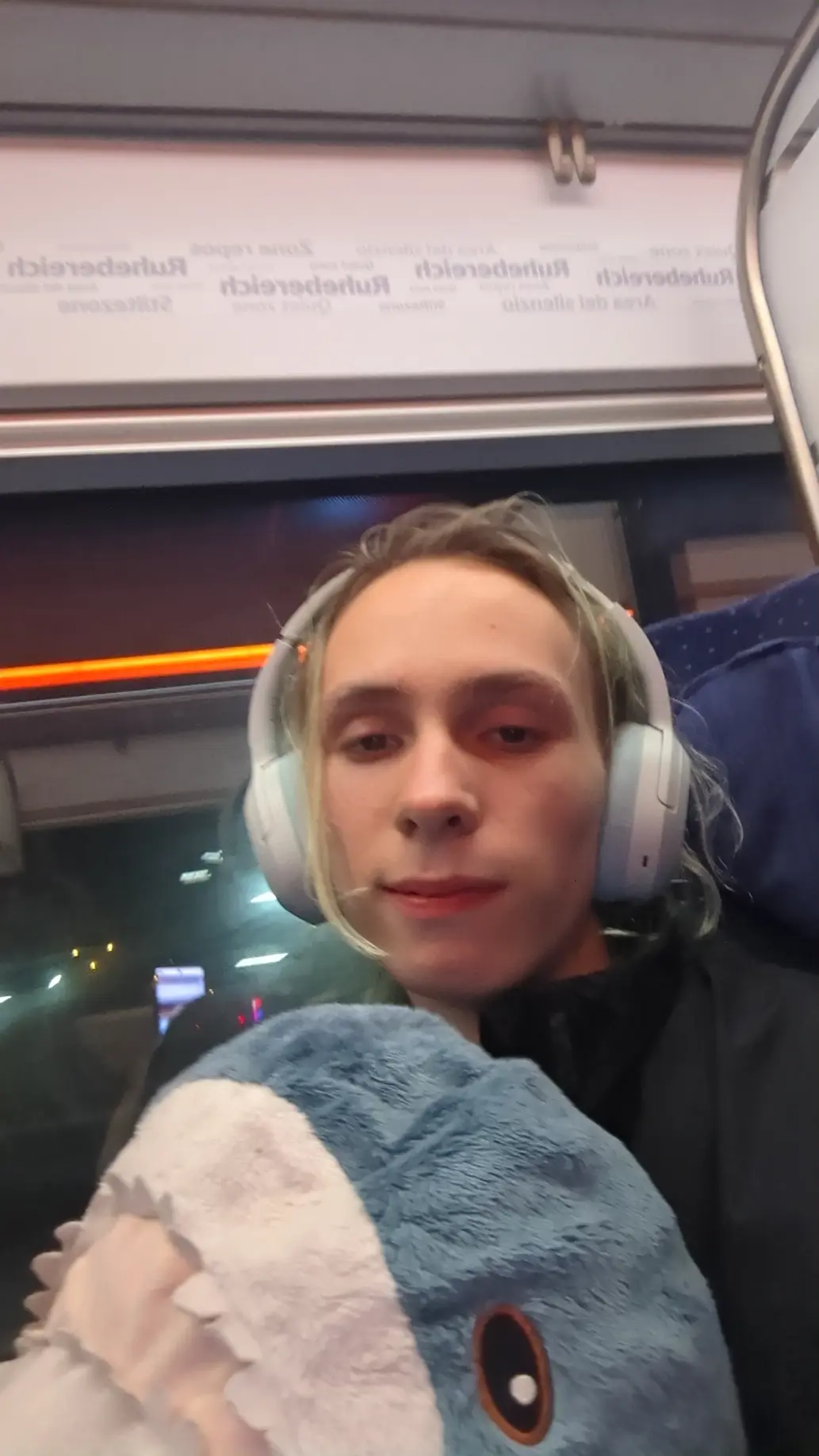 Selfie of me wairing my blue headphones and holding a big blahaj in an ice 4 train with blue seats